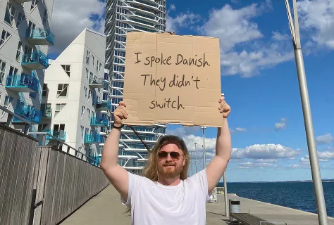 A Swap Language learner on a Danish harbour promenade holding a handwritten cardboard sign reading “I spoke Danish. They didn’t switch.”