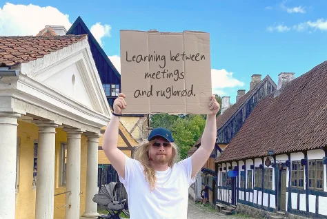 A Swap Language learner in a street of old Danish houses holding a handwritten cardboard sign reading “Learning between meetings and rugbrød.”
