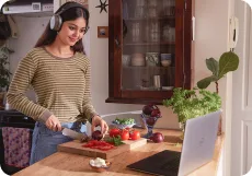 A learner wearing headphones, chopping vegetables at home while a video lesson plays on her laptop